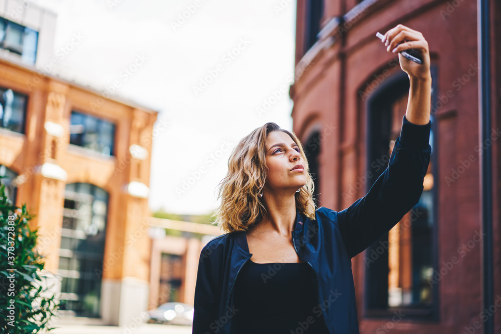 Trendy woman using smartphone for taking selfie on street near buildings