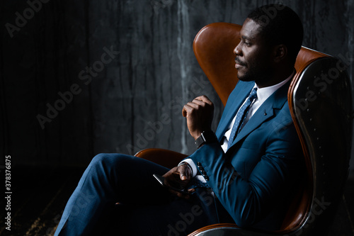 African American businessman in a blue classic suit sits in an aviator loft armchair with a phone in his hands