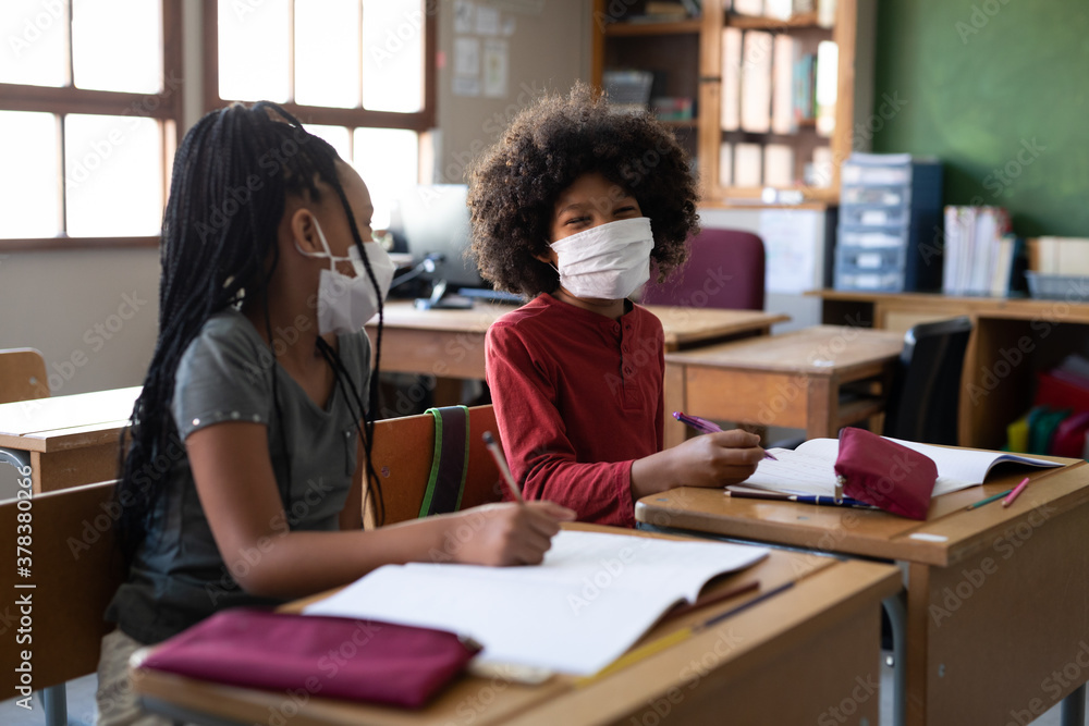 Boy and Girl wearing face masks talking to each other while sitting on their desk at school Boy and Girl wearing face masks talking to each other while sitting on their desk at school