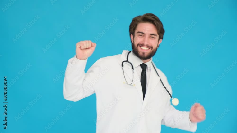 Smiling doctor funny dancing, success and luck at work. Young handsome doc man in professional medical white coat is isolated on blue studio background Smiling doctor funny dancing, success and luck at work. Young handsome doc man in professional medical white coat is isolated on blue studio background