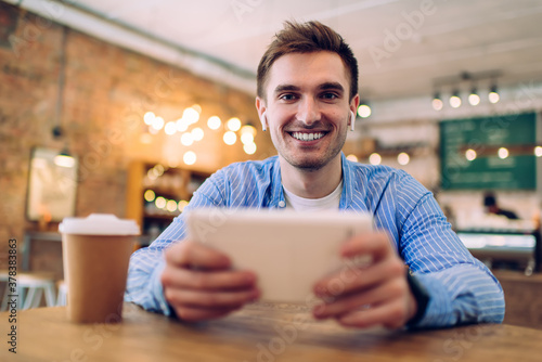 Portrait of cheerful music lover excited with updated audio playlist listening popular tracks and laughing at camera in coffee shop, sincerely man using digital tablet and bluetooth headphones