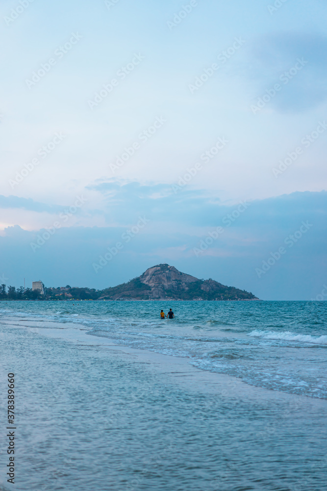 The waves hit the coast in the evening. A woman and a man were Hands in hand together in the middle of ocean. The background has an island with a clear sky in the soft. There is a copy space.