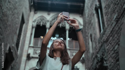 Beautiful long-haired brunette making photo under the arch. Young attractive woman taking pictures of stunning buildings. Pretty tourist makes selfie in a beautiful city. Girl with mobile phone.