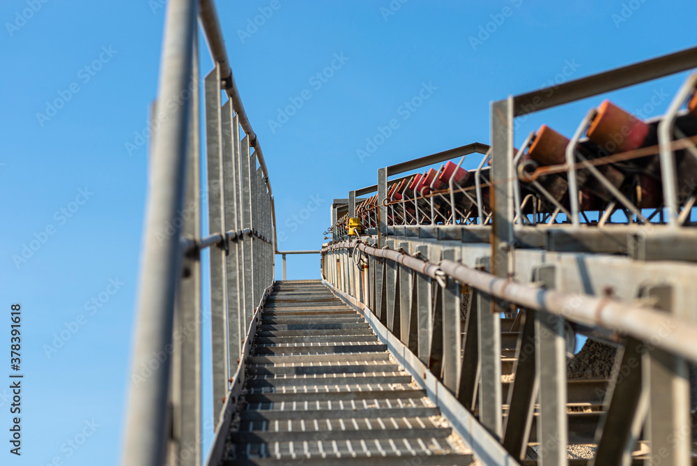 Close-up shot of the conveyor belt in the concrete plant with transport rollers, visible metal stairs and railings. Close-up shot of the conveyor belt in the concrete plant with transport rollers, visible metal stairs and railings.