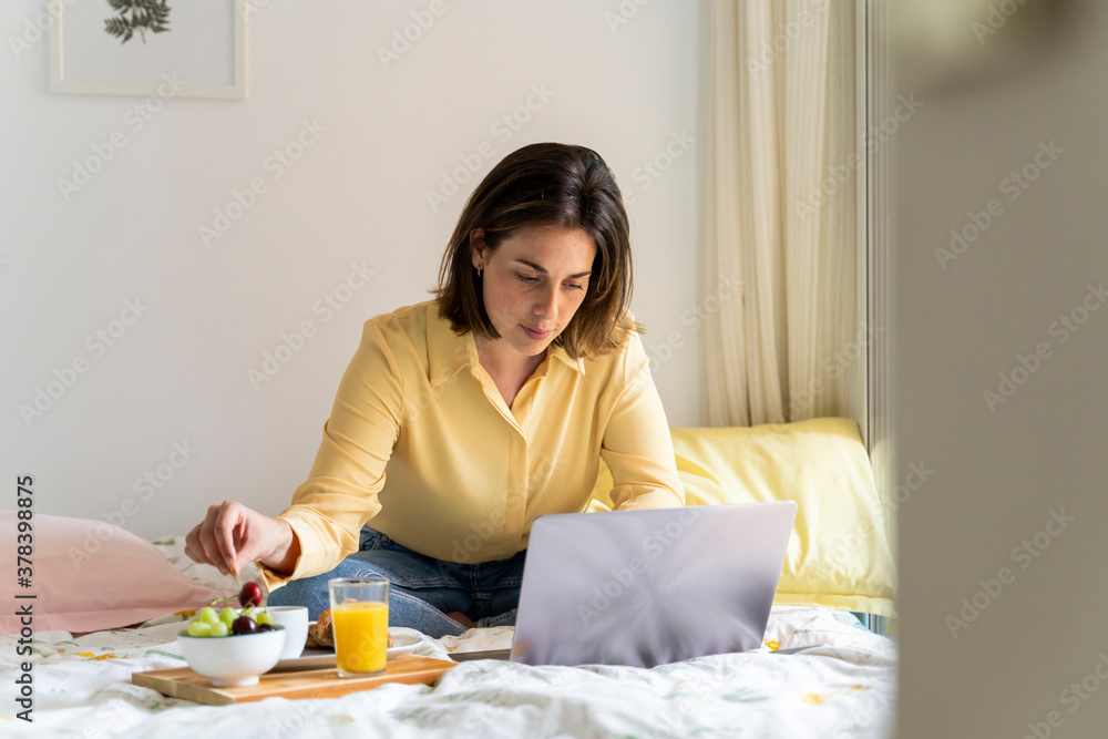 Woman eating breakfast while using laptop in bedroom Woman eating breakfast while using laptop in bedroom