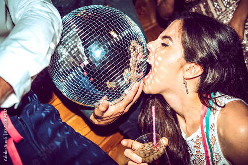Young woman with eyes closed holding drink and biting disco ball in glamorous party photo