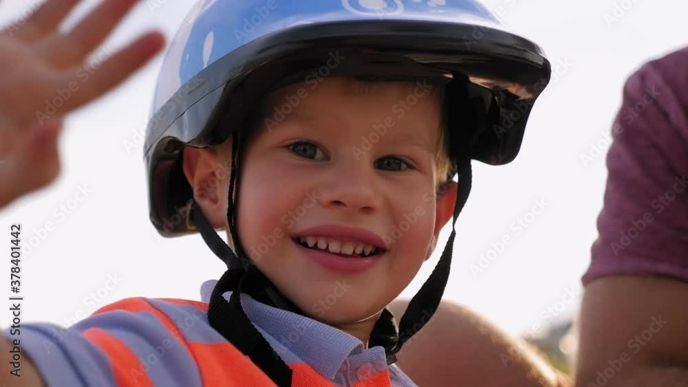 Little boy in a blue bicycle helmet waving his hand while looking at the camera in the summer park. Little boy in a blue bicycle helmet waving his hand while looking at the camera in the summer park.