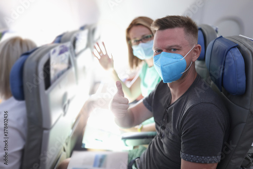 Man with protective mask sit on plane and show his thumbs up. Woman sit next to her husband and wave her hand.