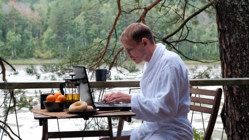 Young Man in a White Bathrobe Using Laptop while Having Breakfast on Vacation. Traveling, Vacation, Remote Work and Freelancer Lifestyle Concept Young Man in a White Bathrobe Using Laptop while Having Breakfast on Vacation. Traveling, Vacation, Remote Work and Freelancer Lifestyle Concept
