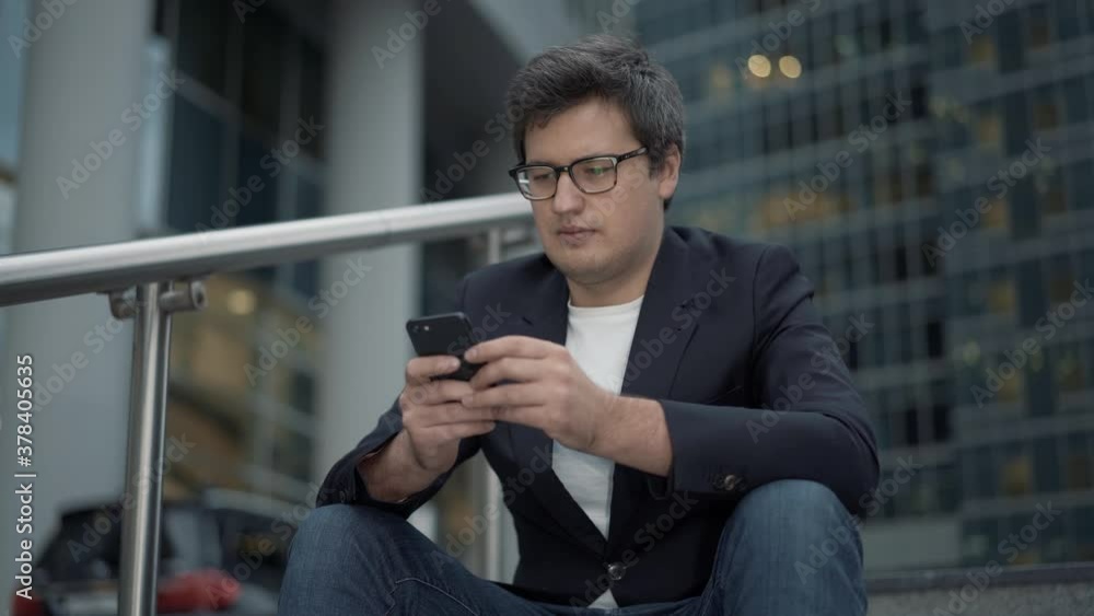 Businessman sitting on stairs on background of glass buildings, windy weather in the evening. Man in black suit alone sitting on the steps with a phone in hand Businessman sitting on stairs on background of glass buildings, windy weather in the evening. Man in black suit alone sitting on the steps with a phone in hand
