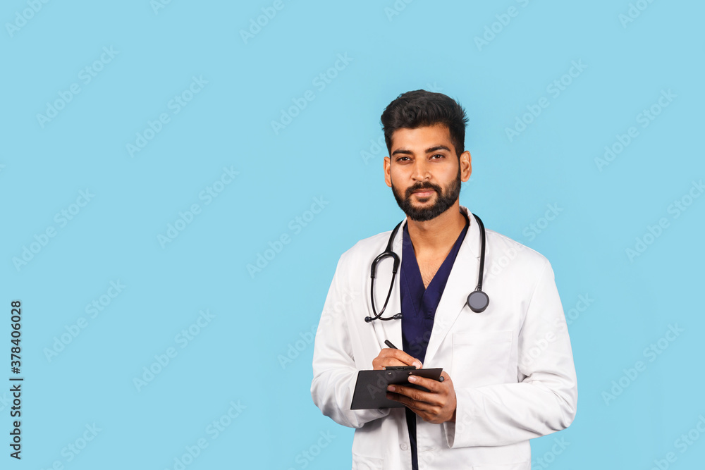 Handsome modern indian / asian doctor with stethoscope, in uniform writing prescription on tablet on blue background Handsome modern indian / asian doctor with stethoscope, in uniform writing prescription on tablet on blue background