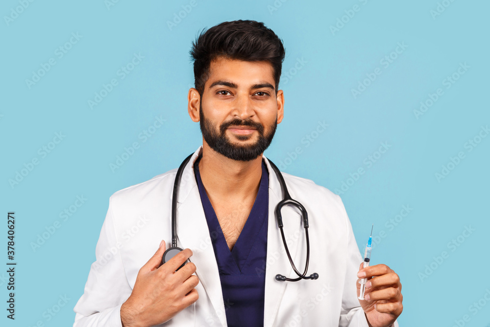 Cute Indian / Asian doctor with a stethoscope and a syringe in his hands, in a white coat on a blue background. Cute Indian / Asian doctor with a stethoscope and a syringe in his hands, in a white coat on a blue background.
