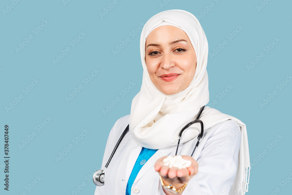 a friendly, Muslim woman doctor in a hijab with a stethoscope holding a handful of pills against a blue background. a friendly, Muslim woman doctor in a hijab with a stethoscope holding a handful of pills against a blue background.