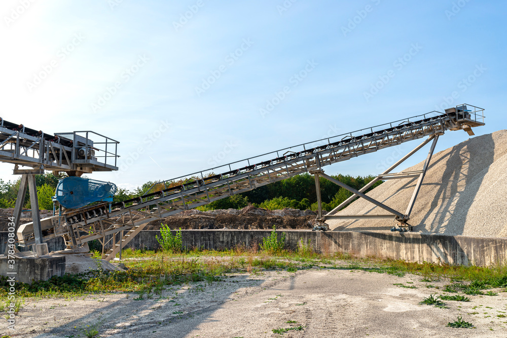 A system of interconnected conveyor belts over heaps of gravel against a blue sky at an industrial cement plant.