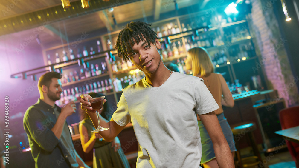 Where party never ends. Cheerful mixed race young man looking at camera, posing with a cocktail in his hand. Friends celebrating, chatting, having fun in the background