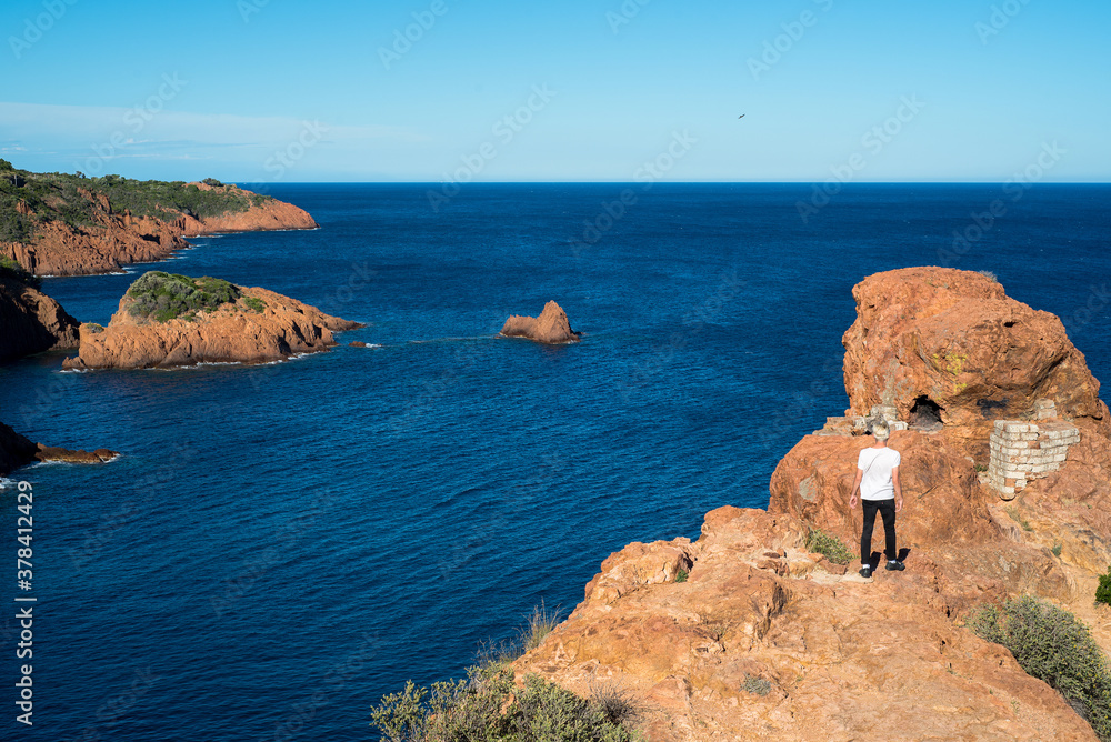 vue panoramique sur une mer bleu azur et une côte de rochers rouges et un marcheur 