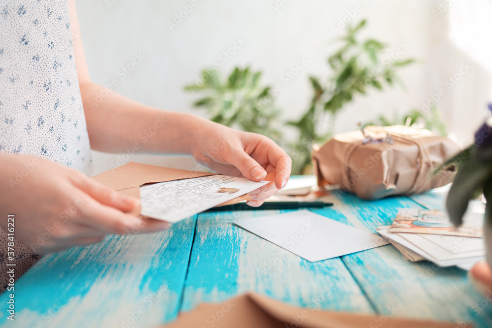 A woman holding a postcard with a craft envelope. Hands close-up. Blue wooden table with a parcel in the background. Concept of postcrossing and communication A woman holding a postcard with a craft envelope. Hands close-up. Blue wooden table with a parcel in the background. Concept of postcrossing and communication