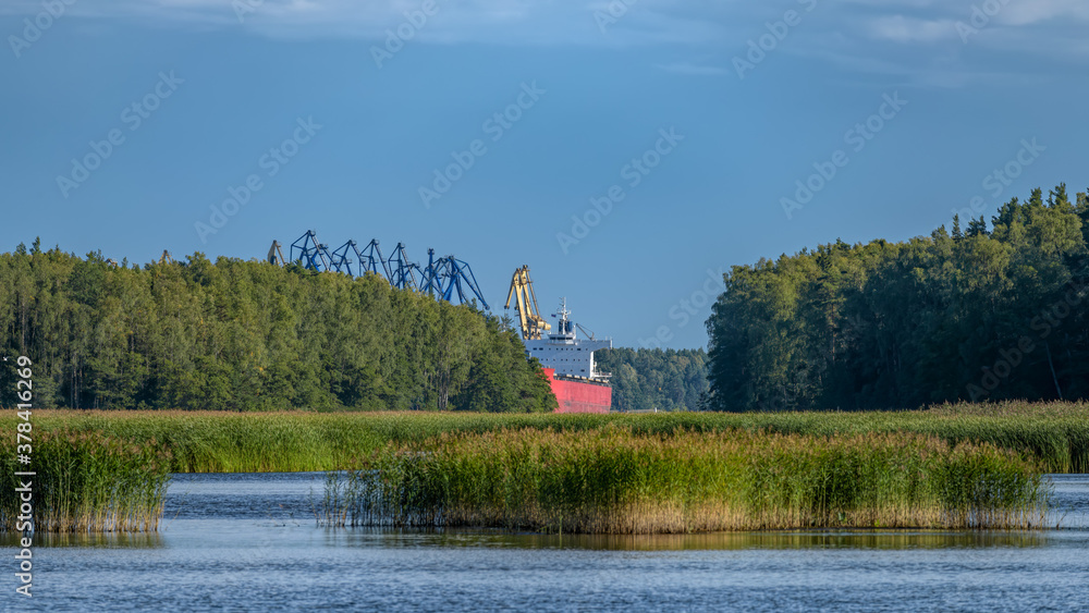 Obraz premium Dry cargo vessel with red-orange hull and gantry cranes of coal handling terminal in Vysotsk cargo port viewed from fairway through strait between islands in Vyborg Bay.
