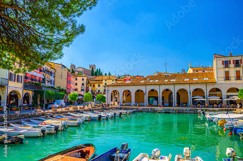 Old harbour Porto Vecchio with motor boats on turquoise water, green trees and traditional buildings in historical centre of Desenzano del Garda town, blue sky background, Lombardy, Northern Italy photo