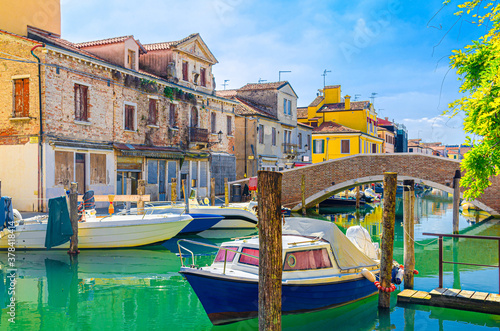 Chioggia cityscape with narrow water canal with moored multicolored boats and yachts between old colorful buildings and brick bridge, blue sky background in summer day, Veneto Region, Northern Italy