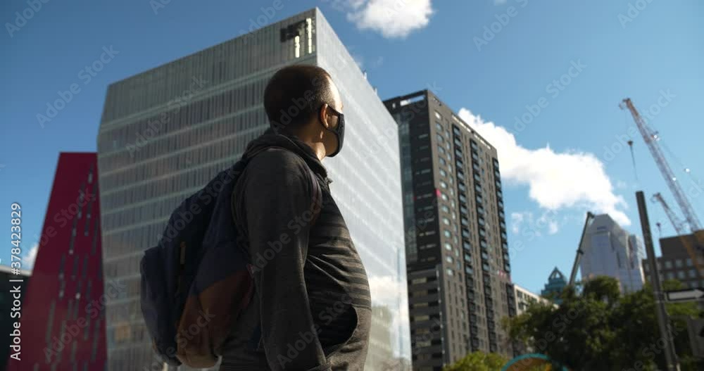 Low angle view of young man wearing covid-19 protective face mask, with backpack moving in city looking around at tall glass building in downtown Low angle view of young man wearing covid-19 protective face mask, with backpack moving in city looking around at tall glass building in downtown