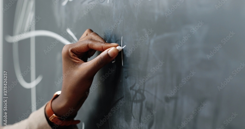 Close up of African American hand of woman writing math or physics formulas with chalks on blackboard. Female teacher at school. Lesson pf mathemathics.