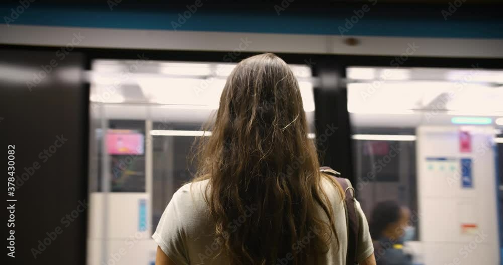Rear view of young woman wearing covid-19 protection face mask waiting at platform of metro station while looking at arriving train Rear view of young woman wearing covid-19 protection face mask waiting at platform of metro station while looking at arriving train