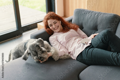 Beautiful redhead woman resting on a sofa laughing and cuddling a dog