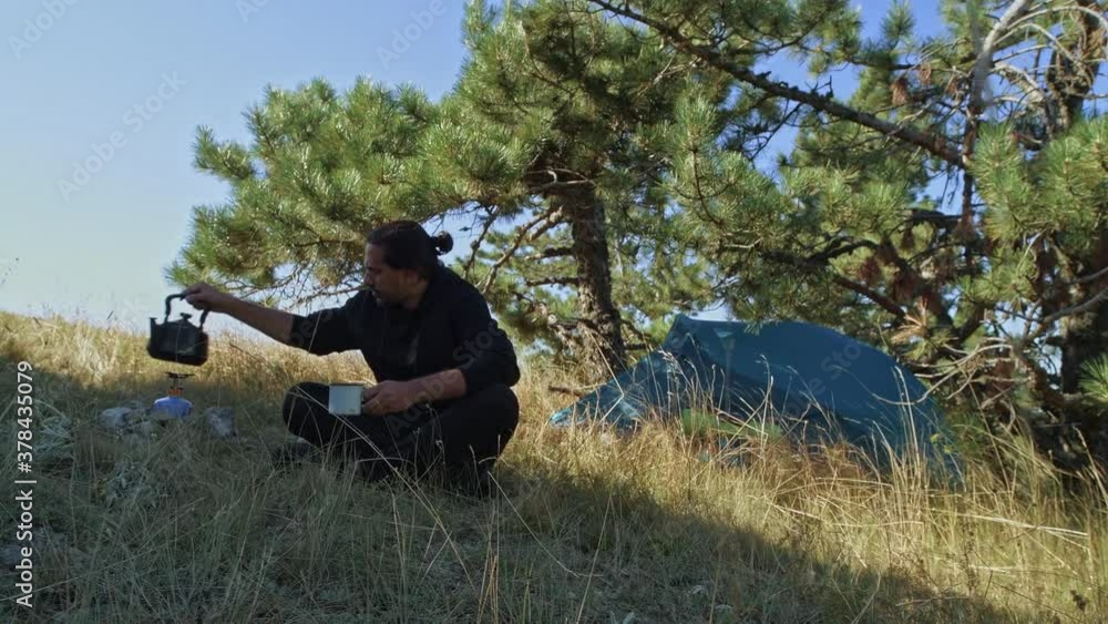 A young, handsome man in a campground light a gas burner. Boiling water. Parking in a tent. A tourist is drinking tea from a cup A young, handsome man in a campground light a gas burner. Boiling water. Parking in a tent. A tourist is drinking tea from a cup