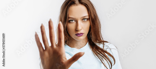 Stop. Enough. Young beautiful girl stretches out her hand to the camera to stop the effect. Woman with fashion makeup with purple lips on an isolated gray background. Bad events, despair and threat.