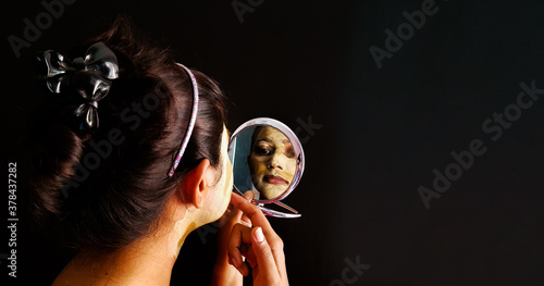 an indian lady with face pack applied in half dried condition checking her face in a mirror with selective focus on the mirror image