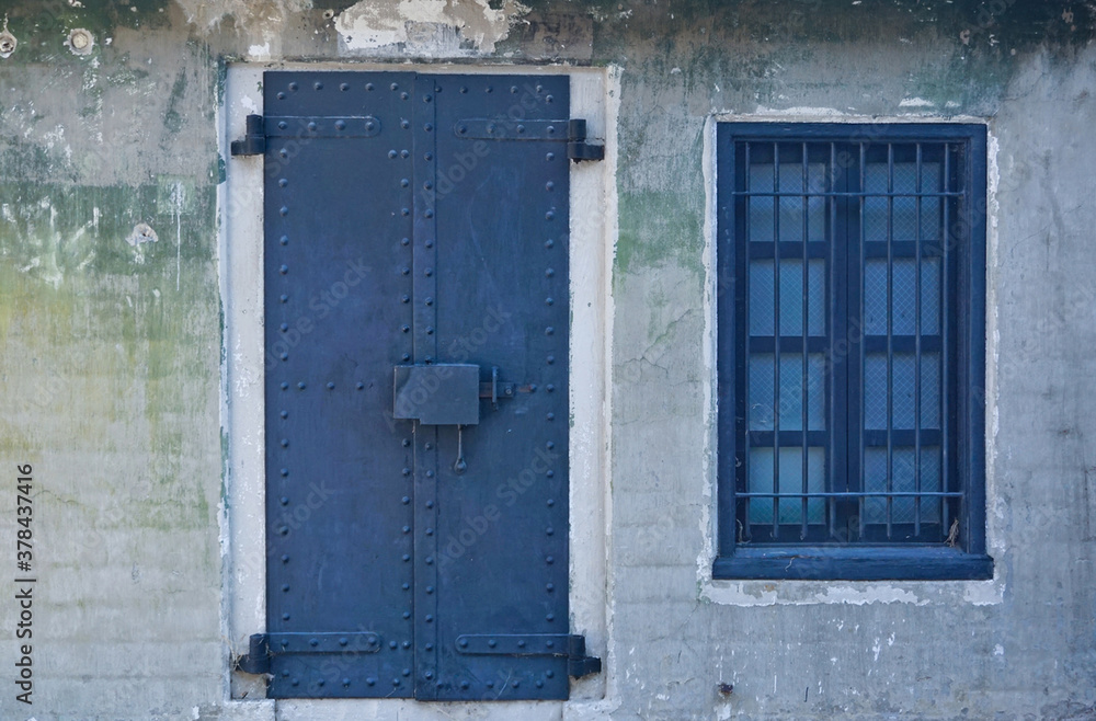 Old stone building front details, black metal door, barred window, textured wall in grays and greens. Old stone building front details, black metal door, barred window, textured wall in grays and greens.