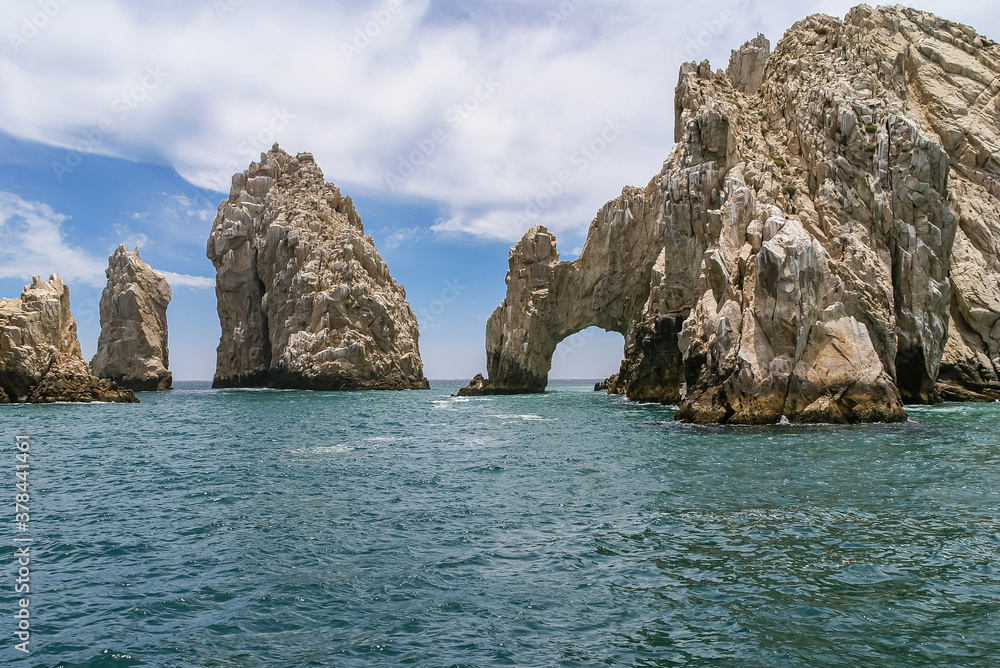Cabo San Lucas, Mexico - April 22, 2008: South end of Baha California. The Arch in beige rocks formation and more free-standing peaks. Greenish ocean water in front under blue cloudscape.
