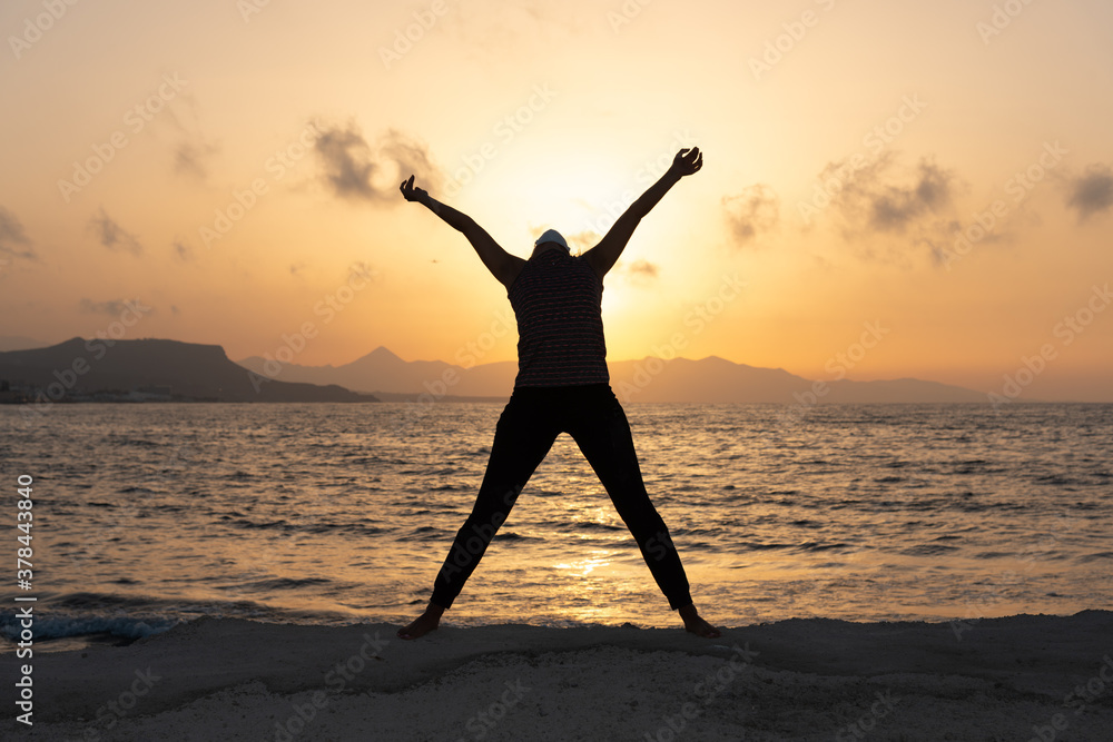 Fototapeta premium Silhouette, young woman with protective surgical face mask performs yoga stretching exercises at the beach at dusk during covid-19 coronavirus pandemic