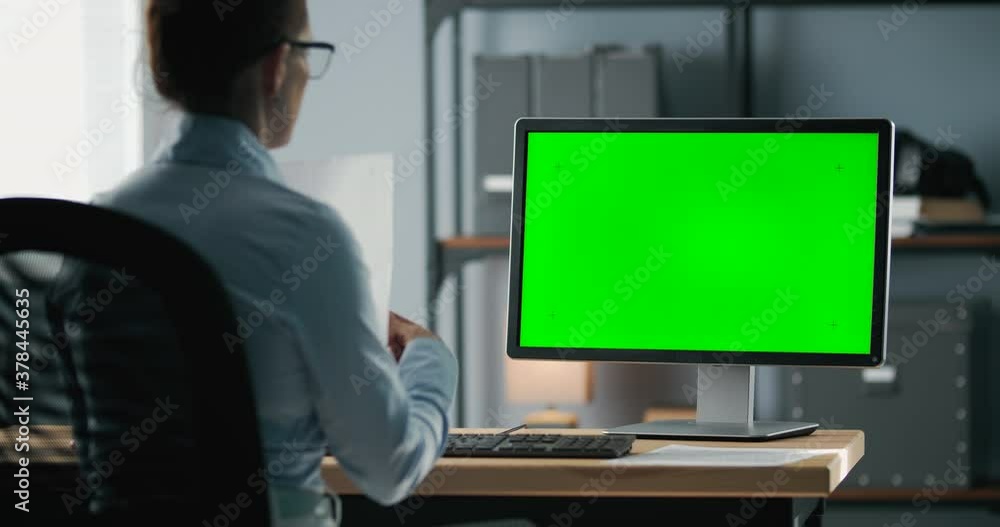 Back view of mature woman in blue shirt talking with colleagues during video chat on computer. Empty chroma key screen. Concept of office work and online conversation. Back view of mature woman in blue shirt talking with colleagues during video chat on computer. Empty chroma key screen. Concept of office work and online conversation.