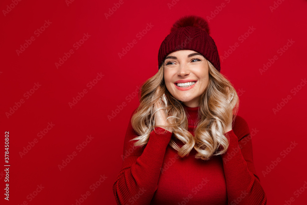 Charming smiling blonde woman touches curly hair with both hands, wears red wool turtleneck and hat, poses against red studio wall, enjoys the winter weather, looking at blank space for advertising Charming smiling blonde woman touches curly hair with both hands, wears red wool turtleneck and hat, poses against red studio wall, enjoys the winter weather, looking at blank space for advertising
