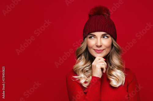 Pretty blonde woman with curly hair touching her chin, wears red wool turtleneck and hat, isolated on red studio wall, enjoys the winter weather, thinking and looking at blank space for advertising