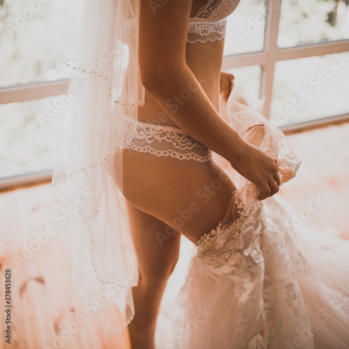 European skinny bride in lingerie puts on a wedding dress. The girl with the veil stuck one leg in the dress.