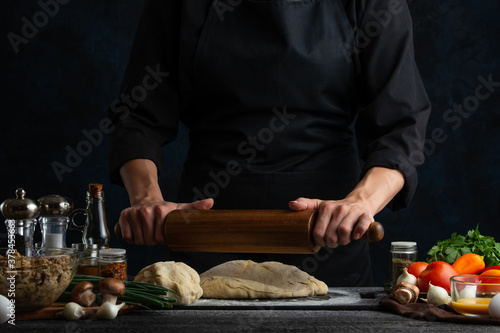 The chef in black uniform with wooden rolling pin in the hands making sweet dough for tasty pie isolated on dark background. Preparing tasty cake. Cooking with love. Homemade dish. Food concept.
