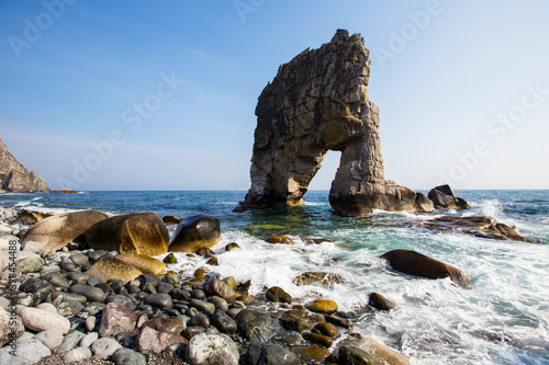 A huge stone in the form of an arch stands in the middle of the sea against the background of sheer cliffs. The territory of the Sikhote-Alin Biosphere Reserve in the Primorsky Territory
