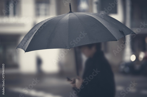 Silhouette with umbrella in rainy foggy day. Focus on umbrella, man in bokeh. Person with umbrella stands by road in rainy weather. Black umbrella, soft selective focus.
