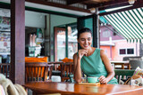Cute elegant calm happy woman in green summer dress sits with coffee in cafe enjoying morning. Beautiful coffee lover. 