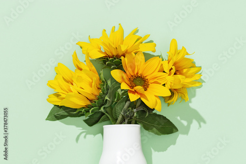 Autumn flower of sunflower in white vase on mint colored background. Natural bright yellow blossom with green leaves.