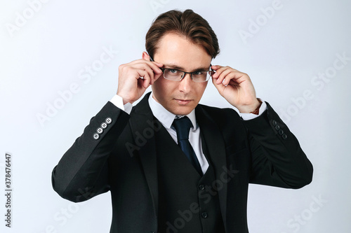A man in a black suit adjusts his glasses. Light background