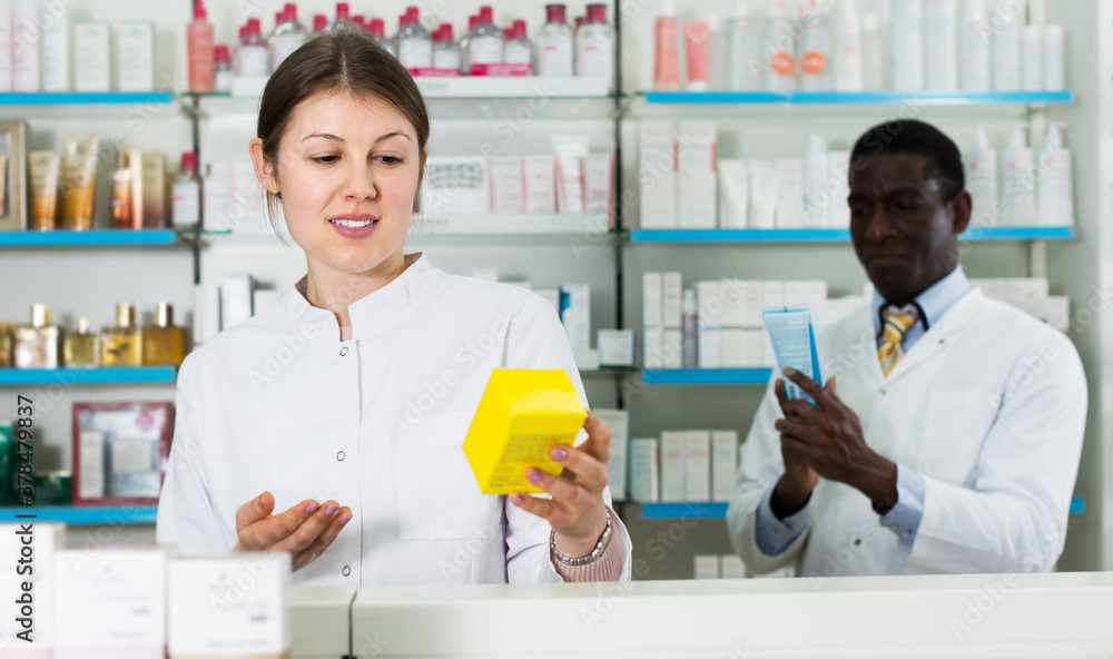 Confident woman pharmacist with medicines on background with male colleague in drugstore