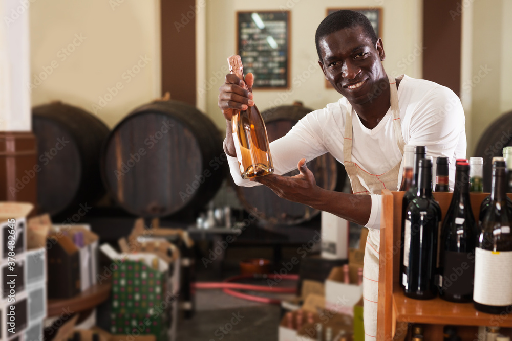 Portrait of cheerful confident afro male seller of wine shop offering wine bottle to buy