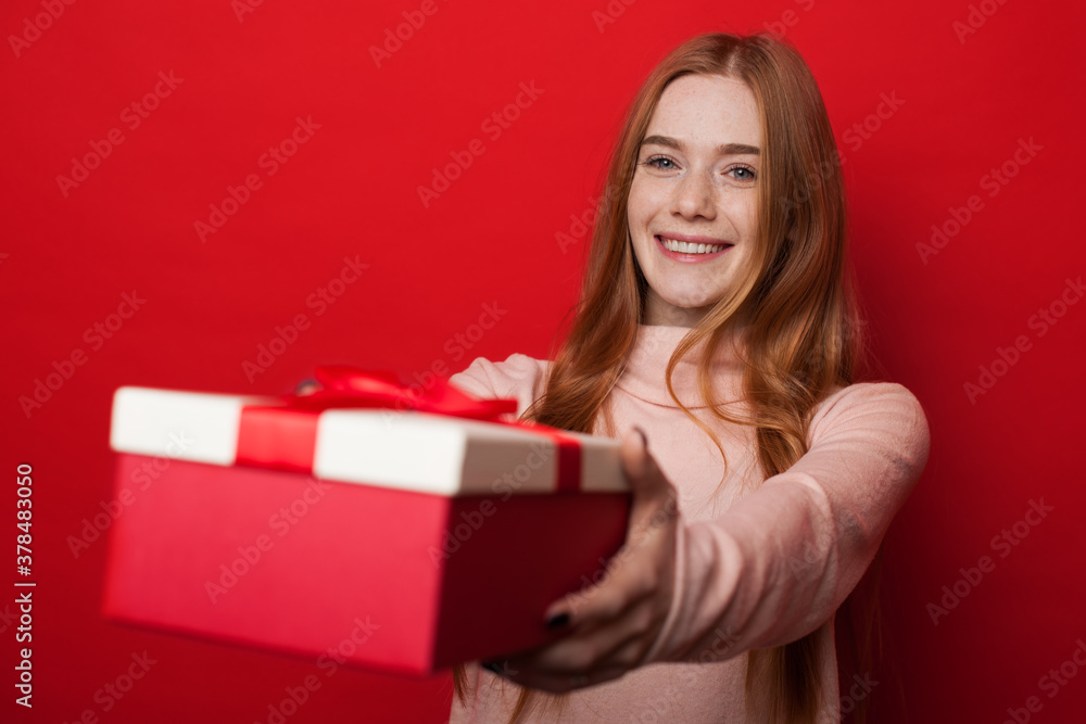 Caucasian woman with freckles and ginger hair is giving a box with gift at camera and smile on red studio wall Caucasian woman with freckles and ginger hair is giving a box with gift at camera and smile on red studio wall