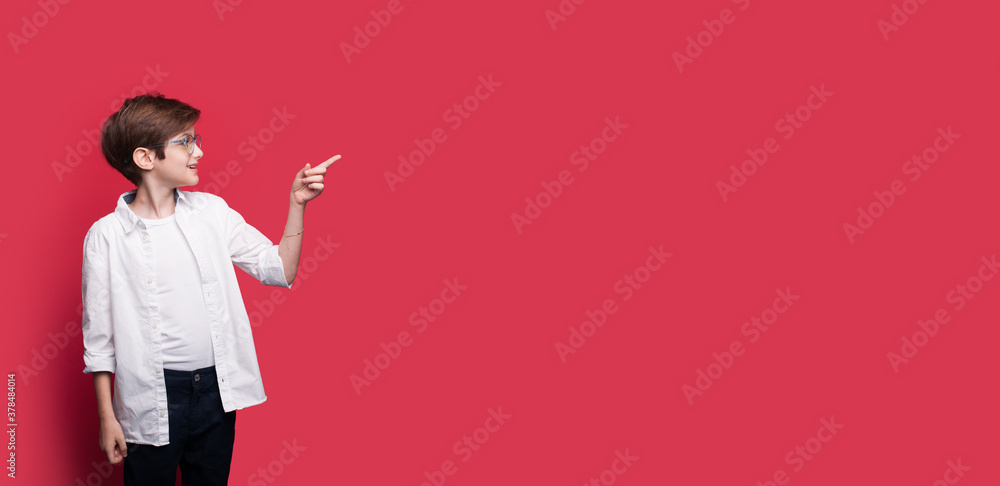 Caucasian boy with glasses and white t-shirt is pointing to red free space near him smiling with opened mouth Caucasian boy with glasses and white t-shirt is pointing to red free space near him smiling with opened mouth
