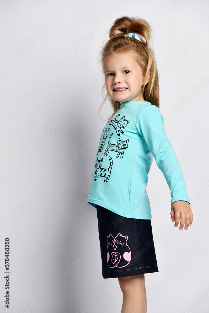 Portrait of smiling blonde kid girl in stylish shirt and denim skirt with cats kitties standing side to camera Portrait of smiling blonde kid girl in stylish shirt and denim skirt with cats kitties standing side to camera