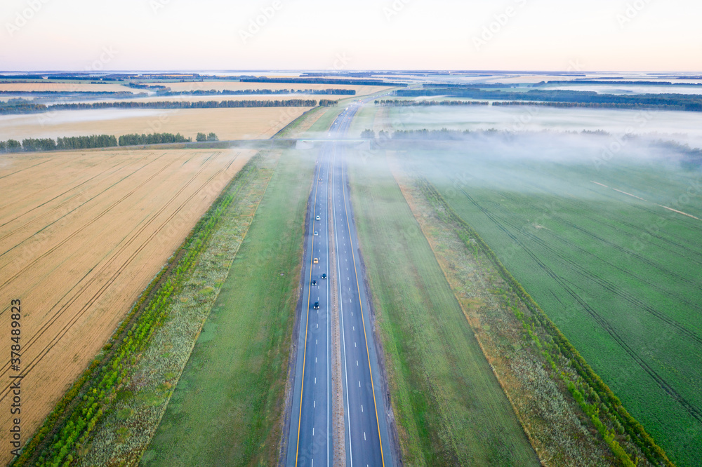 Highway going through meadow in mist surrounded by forest. Aerial view of a road covered in fog.
